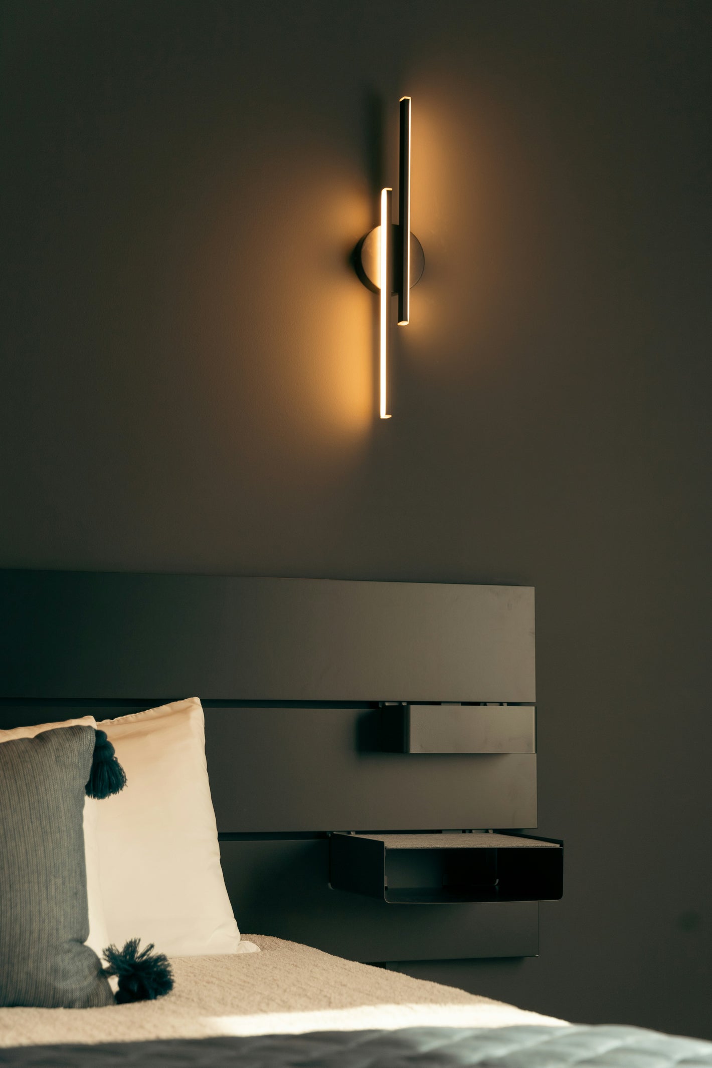 Moderne vertikale LED-Wandleuchte über einem Bett mit dunklem Kopfteil und dekorativen Kissen in einem stilvollen Schlafzimmer.