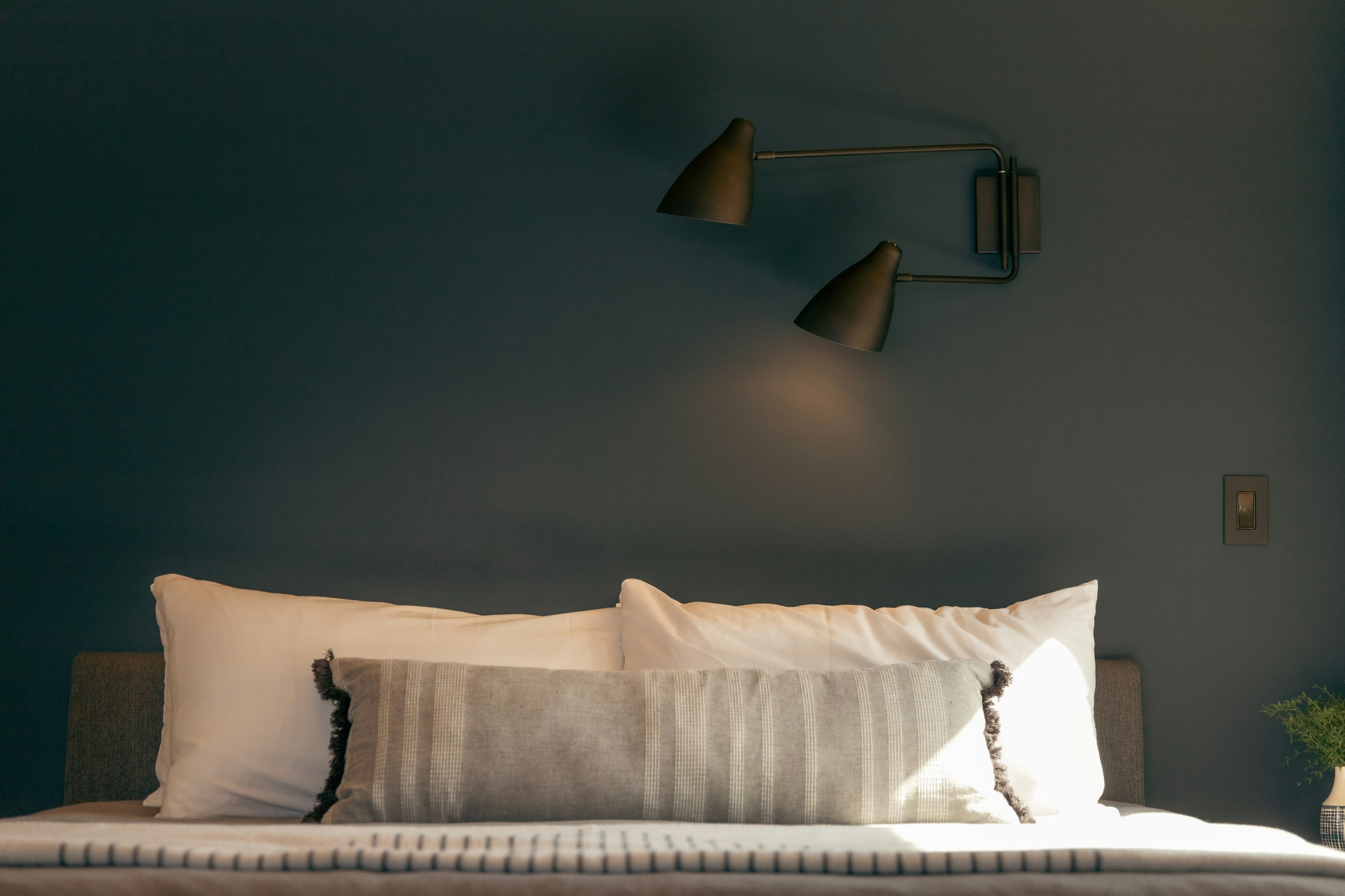 Schlafzimmer mit Doppel-Wandleuchte über Bett mit weißen Kissen und langer Zierkissenrolle vor dunkler Wand.